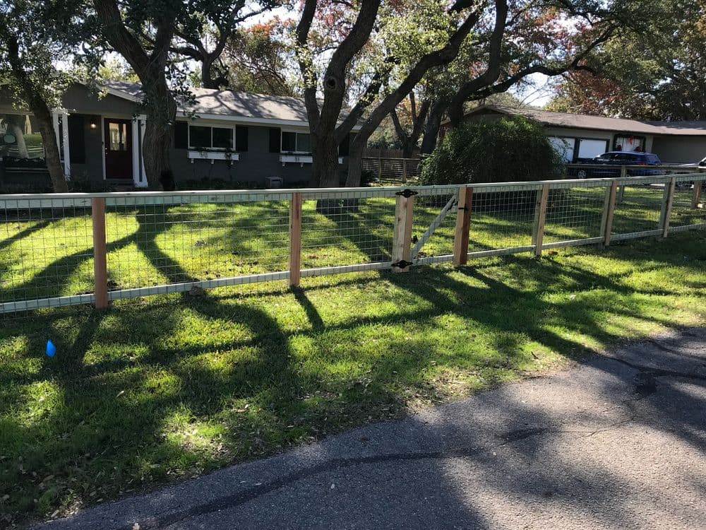 Wooden fence with metal mesh in front of a house surrounded by trees and green lawn.