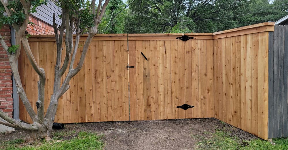 Wooden fence with a gate featuring metal hinges, surrounded by greenery and a brick wall.